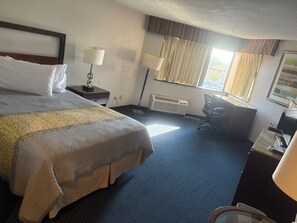 A hotel room with a large bed, a desk, a chair, a lamp, and a window with curtains. - APM Inn and Suites (Hagerstown)