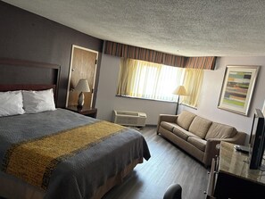 A hotel room with a bed, a sofa, a desk with a computer, a lamp, and a window with curtains. - APM Inn and Suites (Hagerstown)