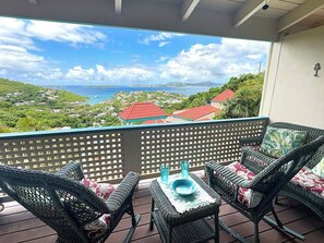 A balcony with wicker furniture and a table, overlooking a coastal landscape with houses and greenery.