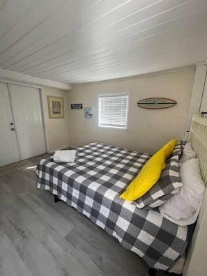 A bedroom with a checkered bedspread, a white radiator, a window with blinds, and a door. - Pet-Friendly 2BR /2 min Walk to Beach /Pool+Hot Tub/ Golf Cart option (North Myrtle Beach)