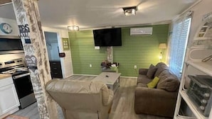 A compact living space with a sofa, a small table, a TV mounted on the wall, and a kitchen area visible in the background. - Pet-Friendly 2BR /2 min Walk to Beach /Pool+Hot Tub/ Golf Cart option (North Myrtle Beach)