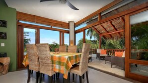 A dining area with a table set for four, surrounded by wicker chairs, a ceiling fan, and large windows offering a view of the outdoors.