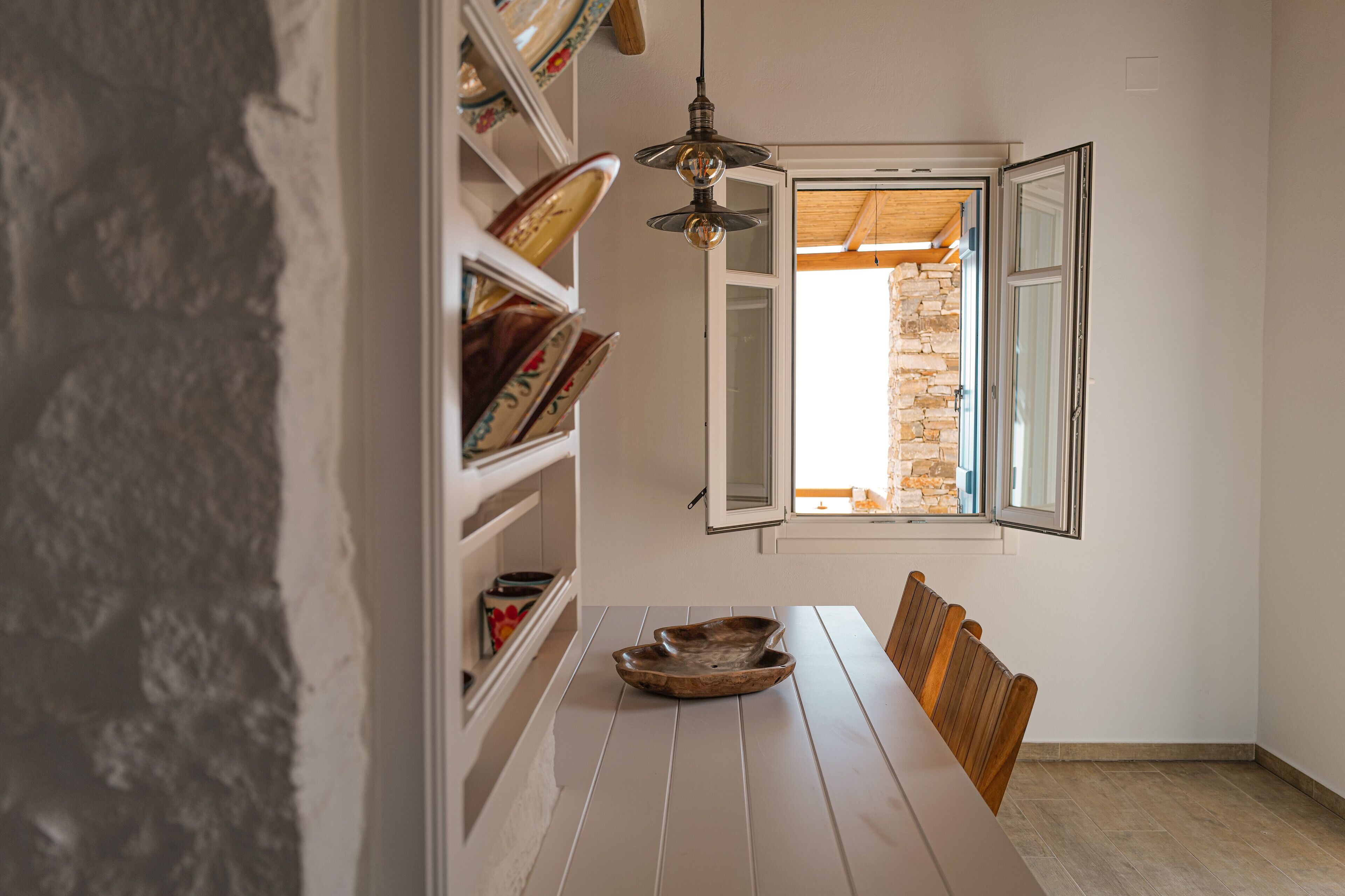 Un coin repas avec une table en bois, des chaises, un luminaire suspendu et une fenêtre donnant sur un mur en pierre.