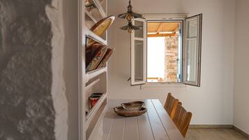 Un coin repas avec une table en bois, des chaises, un luminaire suspendu et une fenêtre donnant sur un mur en pierre.