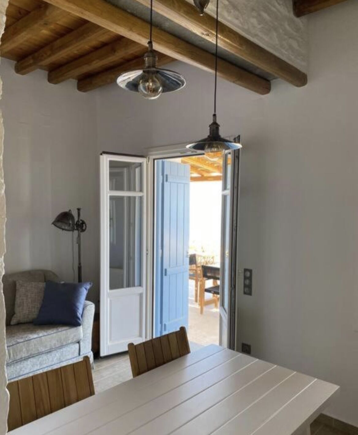 Un coin repas avec une table et des chaises blanches, une porte bleue donnant sur l’extérieur et un plafond avec des poutres en bois apparentes.