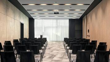 Ett konferensrum med rader av svarta stolar, en väggpanel i trä och ett stort fönster med tunna, genomskinliga gardiner.