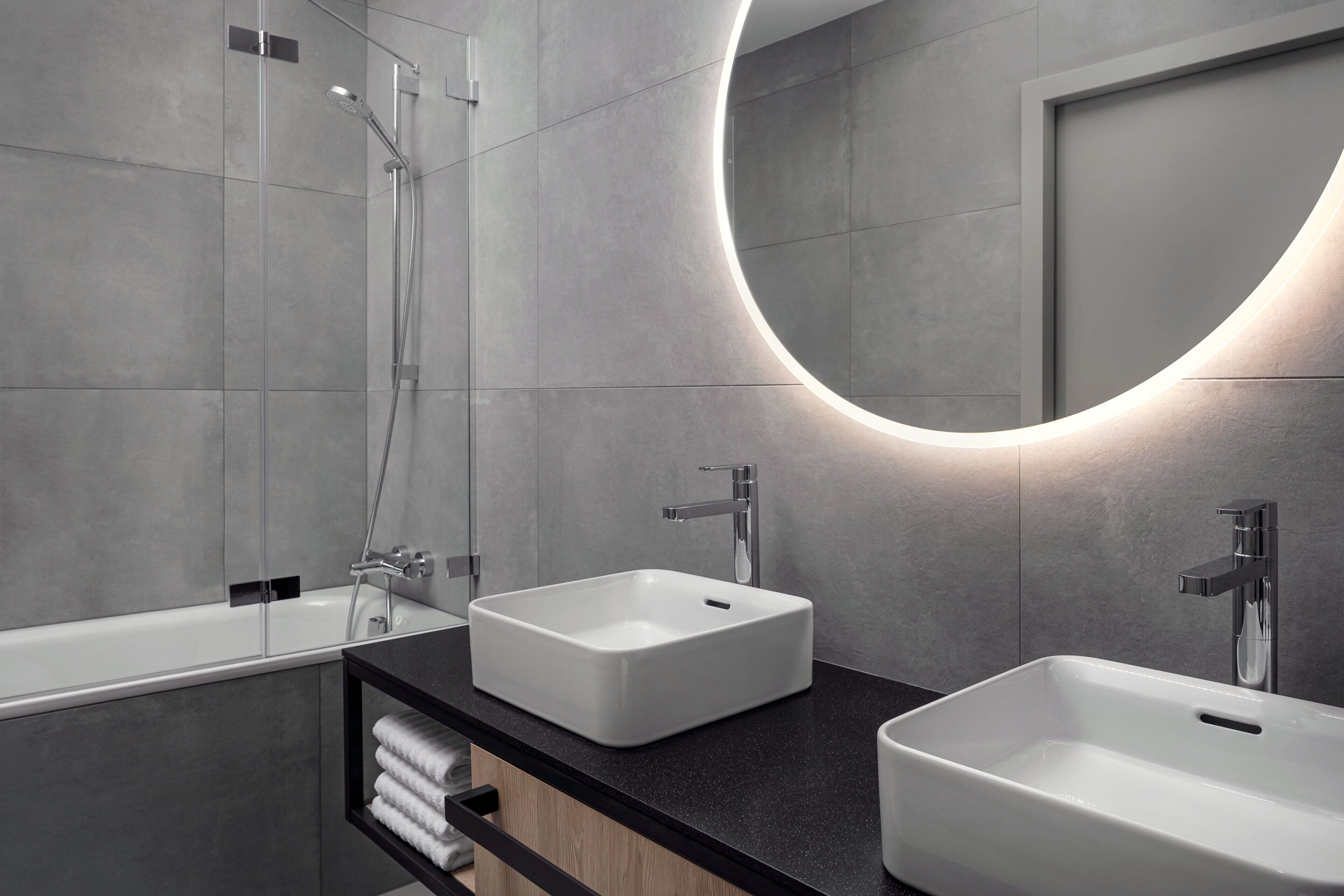 Une salle de bain moderne avec deux lavabos, une douche et un grand miroir agrémenté d’un luminaire circulaire.