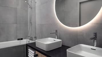 Une salle de bain moderne avec deux lavabos, une douche et un grand miroir agrémenté d’un luminaire circulaire.