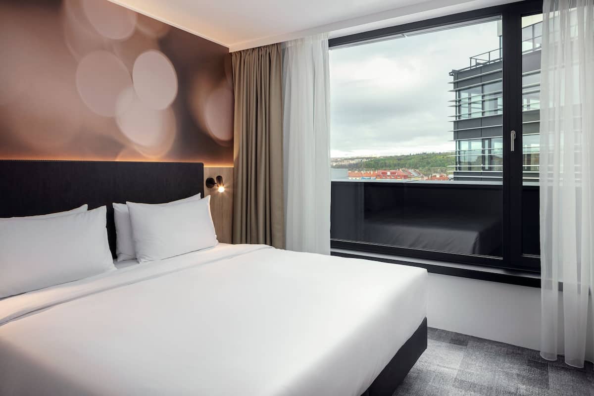 Een moderne hotelkamer met een groot bed, uitzicht op een stadsbeeld door een groot raam en een balkon met stadszicht.