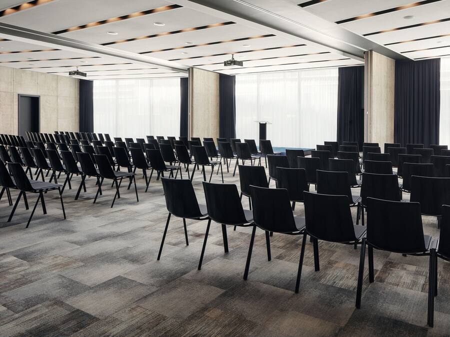Uma sala de conferências com fileiras de cadeiras pretas, um projetor e uma janela grande com cortinas.