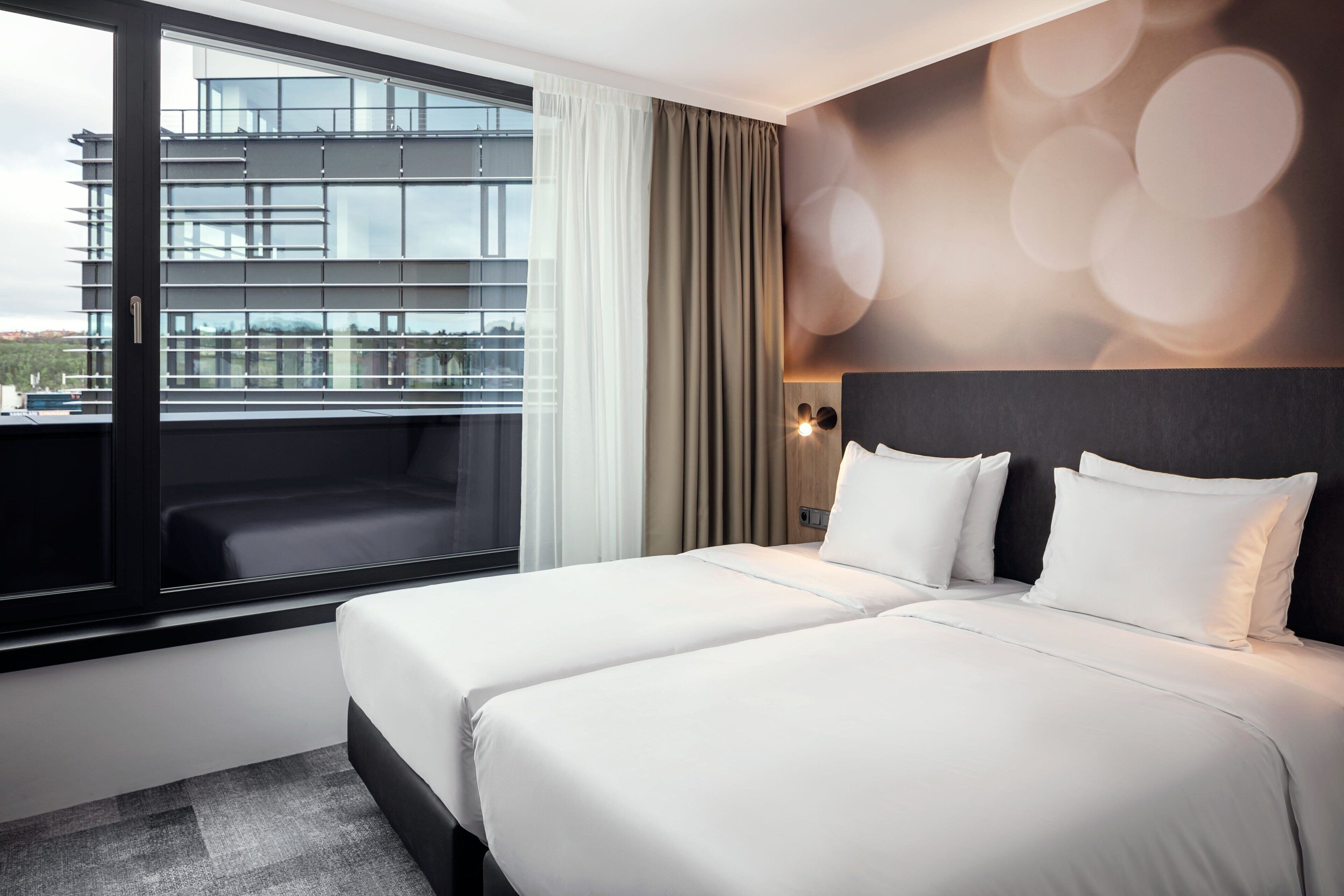A hotel room with two beds, a large window with a view of a building, a dark headboard, and a wall with circular light patterns.