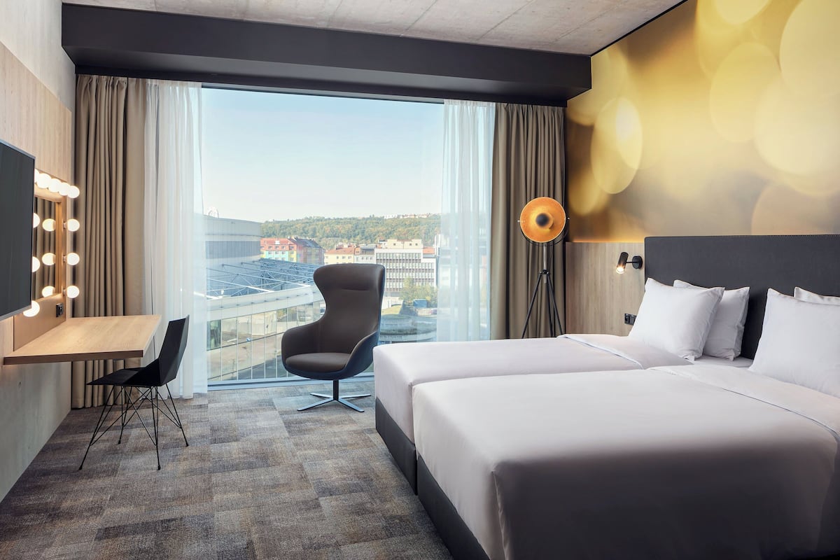 Een hotelkamer met een groot bed, een bureau met stoel, een kaptafel met verlichting, een vloerlamp en uitzicht op de stad door grote ramen.