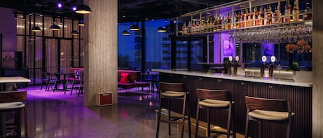 Zona de bar con taburetes altos, una barra y estantes llenos de botellas y vasos.