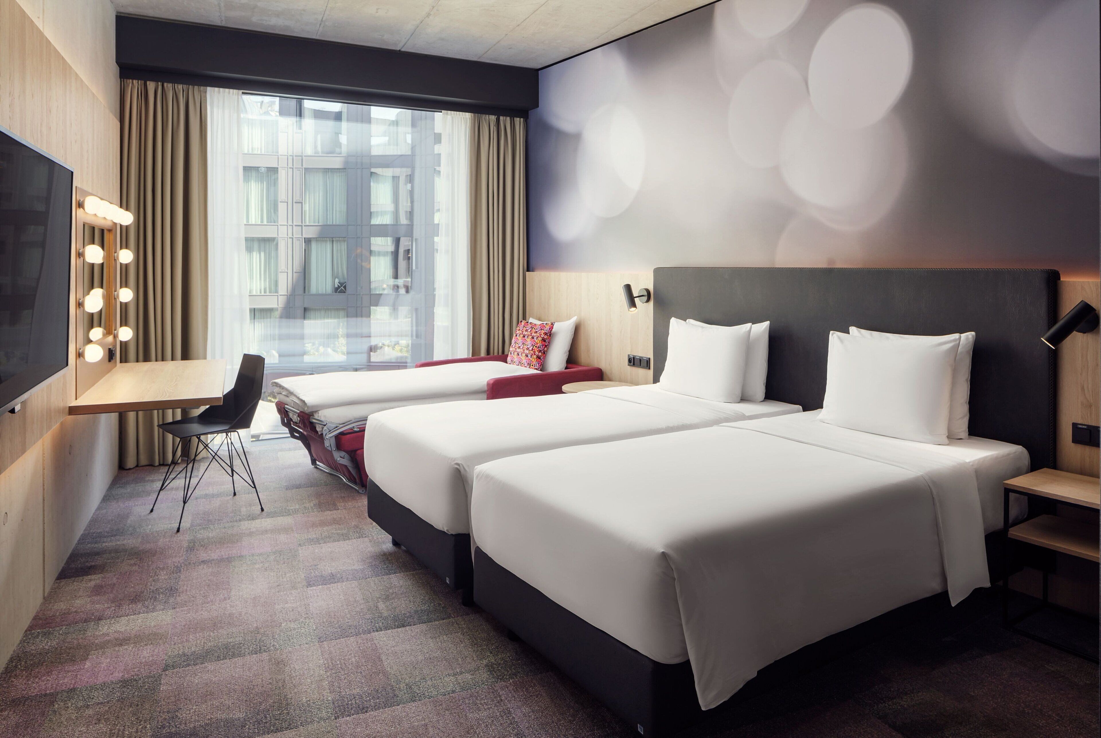 A hotel room with two beds, a desk with a chair, a vanity with lights, and a large window with curtains.