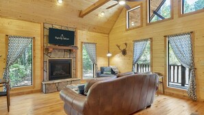 Un acogedor interior de cabaña con chimenea, sofá de cuero, piso de madera y ventanas amplias.