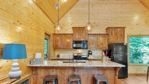 Una cocina estilo cabaña con paredes de madera, una isla central con taburetes, microondas, refrigerador y lámparas colgantes.
