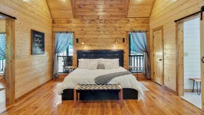 Un dormitorio de cabaña de madera con una cama grande, dos lámparas de noche y vistas al exterior a través de cortinas.