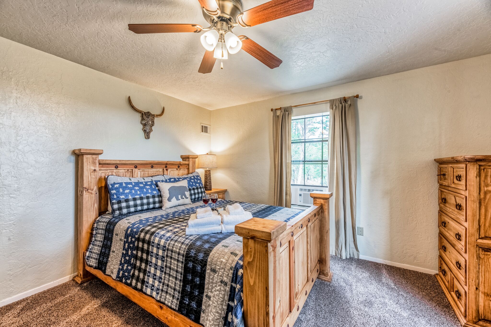 A bedroom with a wooden bed, a ceiling fan, a bull skull decoration, a window with curtains, and a wooden dresser.