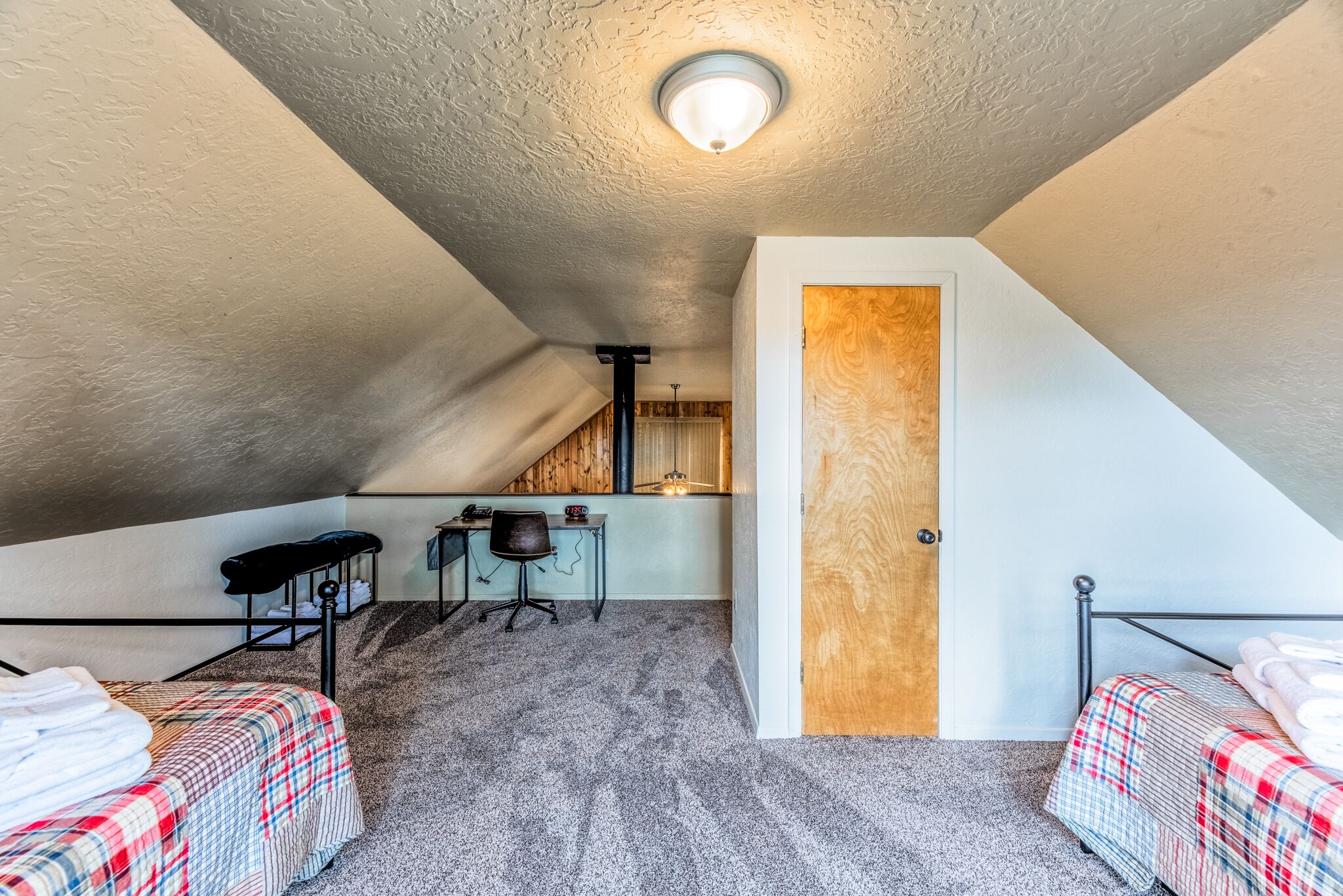 A small, compact room with a sloped ceiling, a kitchenette, two beds with plaid bedding, and a single chair.