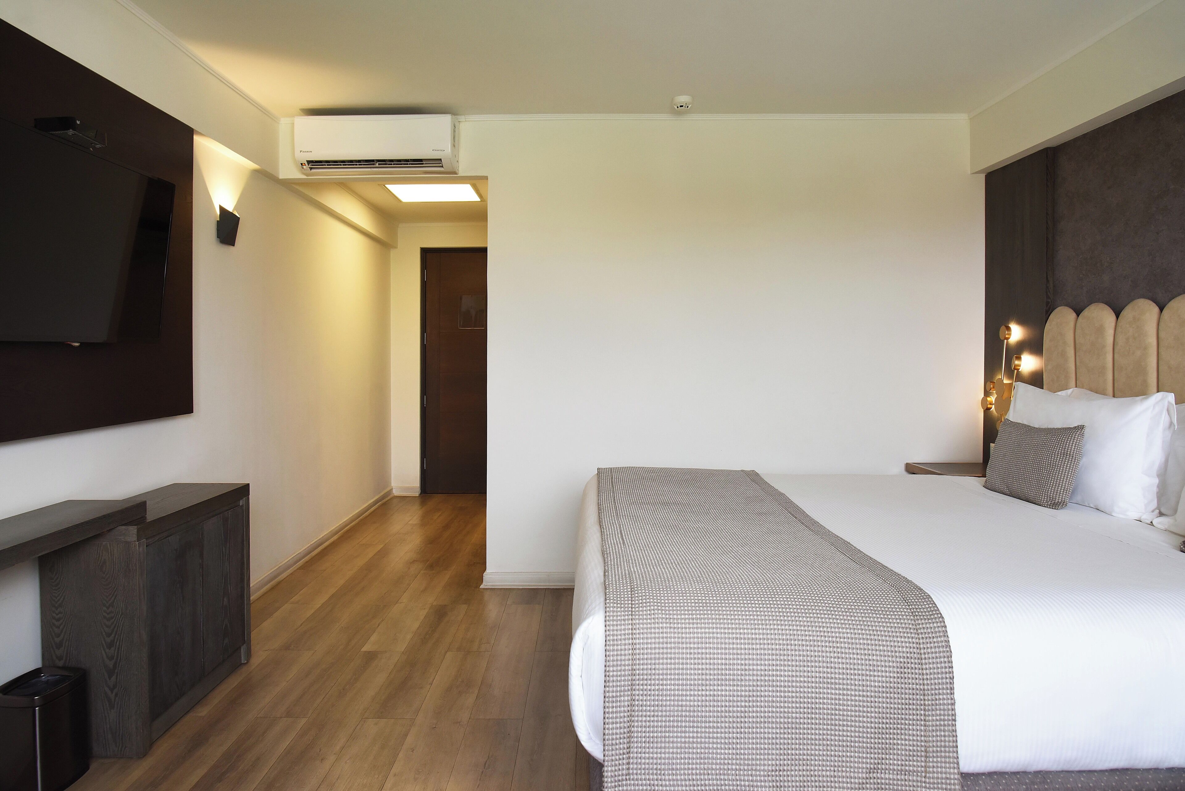 A hotel room with a bed, a TV mounted on the wall, a wooden floor, and an air conditioning unit.