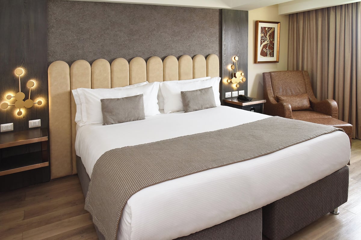 A modern hotel room with a large bed, a brown upholstered armchair, a bedside table with a lamp, and a wall-mounted light fixture.