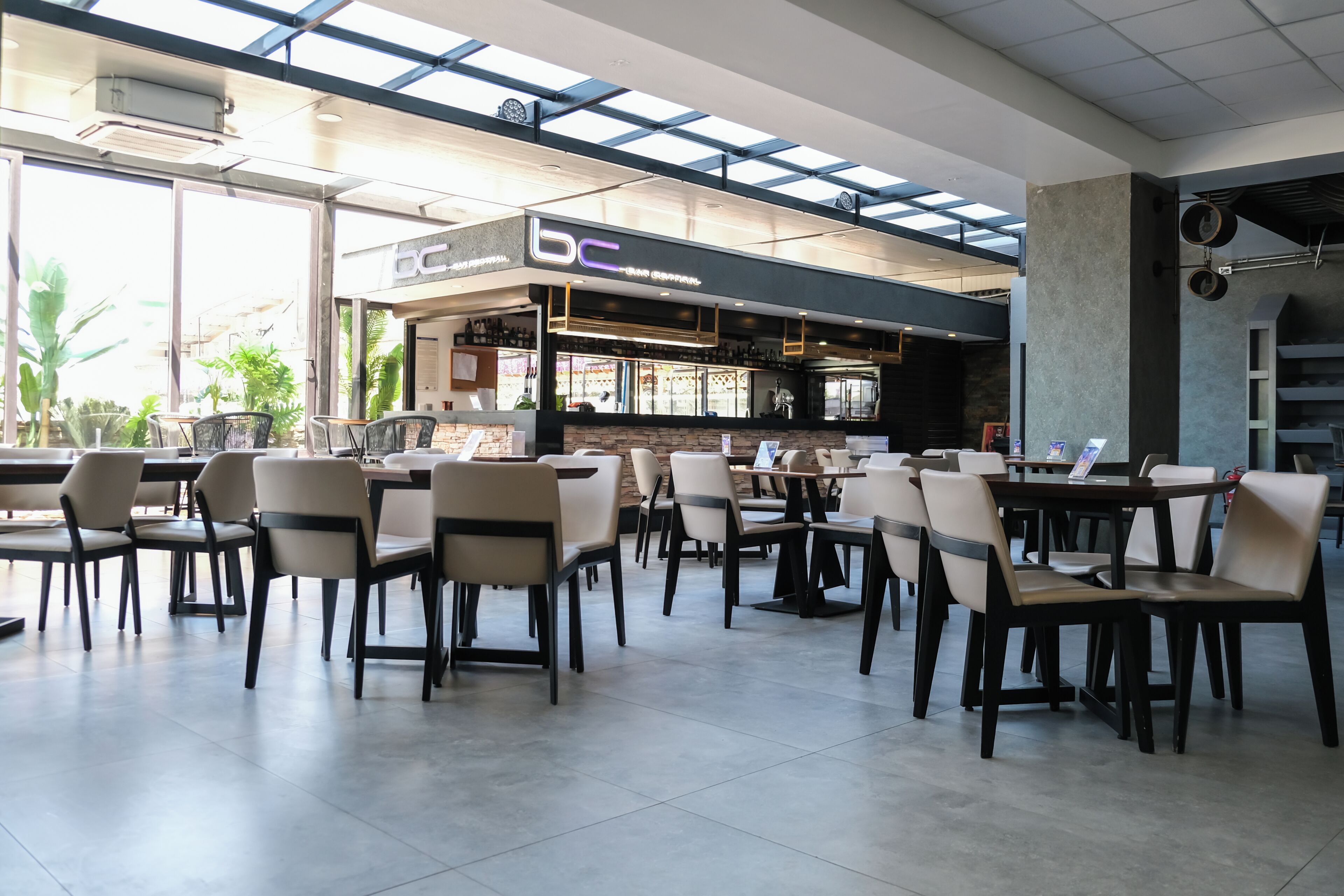 A spacious indoor dining area with multiple tables and chairs, a bar area with a 'bc' sign, and large windows allowing natural light.