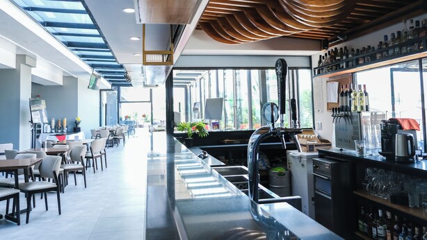 A modern bar area with a long counter, seating on both sides, and a view through large windows. - Hotel HW Libertad (Santiago)