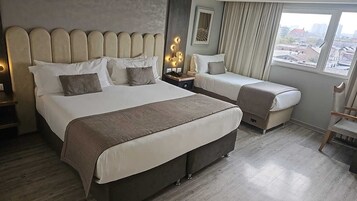 Habitación de hotel con una cama grande, otra más pequeña, un escritorio con lámpara y vistas a la ciudad desde la ventana.