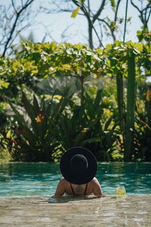A person in a swimming pool with a large black hat, surrounded by lush greenery.