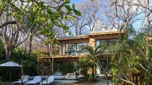 A modern house with a glass facade, surrounded by a garden with lounge chairs and palm trees. -  Luxury Beachfront Villa with private pool at Nantipa (Cóbano)