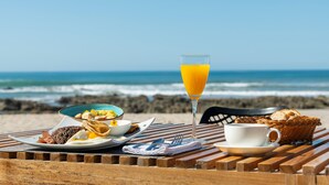 A wooden table with breakfast items including a glass of orange juice, a cup, a plate with food, and a basket of bread on a beach with the ocean in the background. -  Luxury Beachfront Villa with private pool at Nantipa (Cóbano)