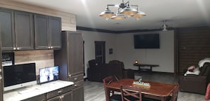 Cozinha moderna com armários escuros, bancada de madeira e televisão de tela plana. Há uma área de jantar com mesa e cadeiras de madeira, e um sofá com um cachorro.