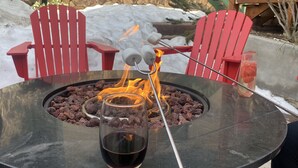Una mesa redonda con una copa de vino tinto, un brasero con leña encendida y dos sillas rojas.