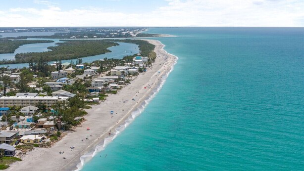 Vista aérea de uma área costeira com uma extensa praia de areia, mar azul cristalino e um conjunto denso de edifícios ao longo da costa.