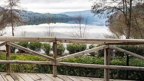 Terraza de madera con vista a un lago, donde se ven casas y árboles a lo lejos.