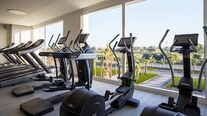 Un gimnasio con varias cintas de correr y máquinas elípticas, amplias ventanas con vistas a un área ajardinada.