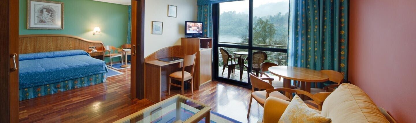 A room with a bed, a desk, a chair, a table, and a balcony with a view.