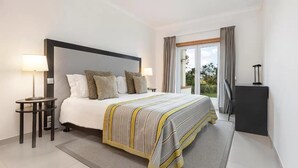 A modern hotel room with a large bed, bedside lamps, a small table, a view of the outdoors through a window, and a dark-colored cabinet. - Vale Da Ribeira Residences (Portimao)