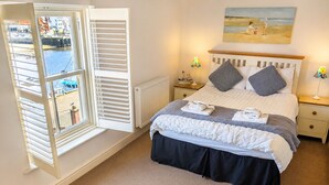 A bedroom with a bed, a window with shutters, a nightstand, a lamp, and a painting on the wall. - Whitby Sea View Cottage Harbour Views Quiet Cragg (Whitby)