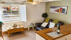 A cozy living room with a wooden coffee table, a leather sofa, a small side table, a window with shutters, and a painting on the wall. - Whitby Sea View Cottage Harbour Views Quiet Cragg (Whitby)