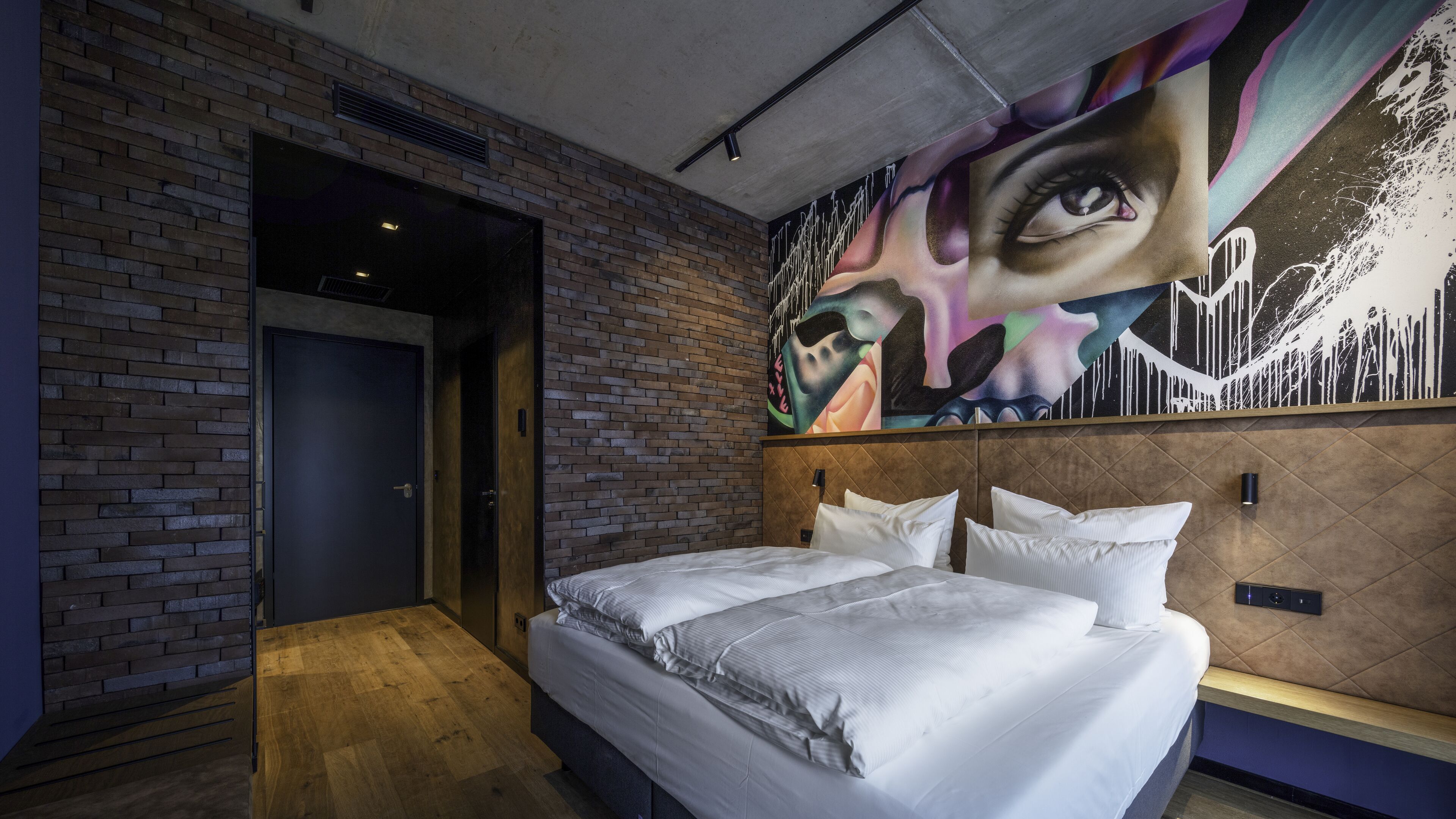 Una habitación de hotel moderna con pared de ladrillo, una cama grande con sábanas blancas, un cabecero con un mural de un paisaje urbano y suelo de madera.