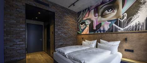 Una habitación de hotel moderna con pared de ladrillo, una cama grande con sábanas blancas, un cabecero con un mural de un paisaje urbano y suelo de madera.