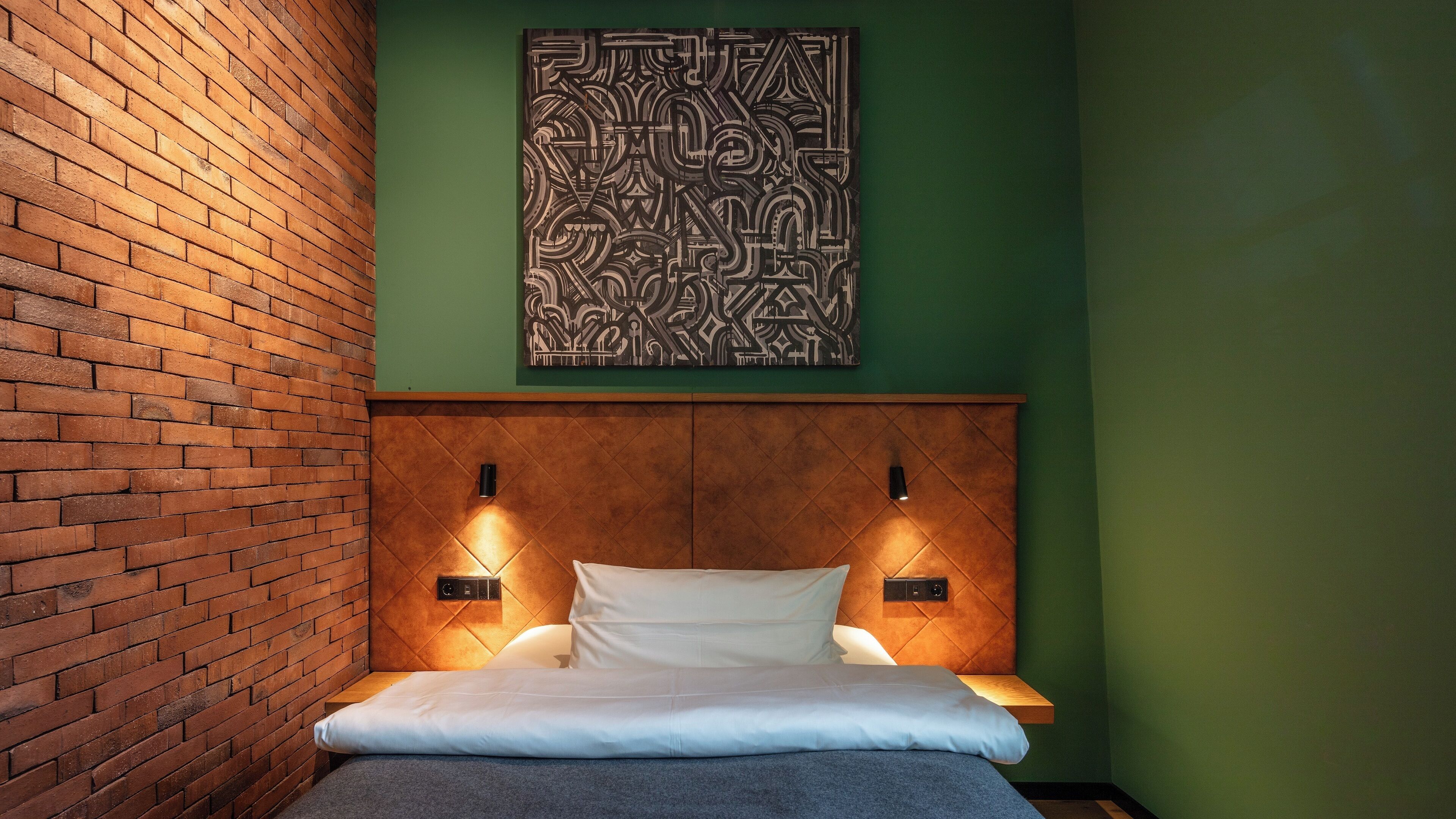 A room with a brick wall, a bed with white bedding, a geometric wall art piece, and two wall-mounted light fixtures.
