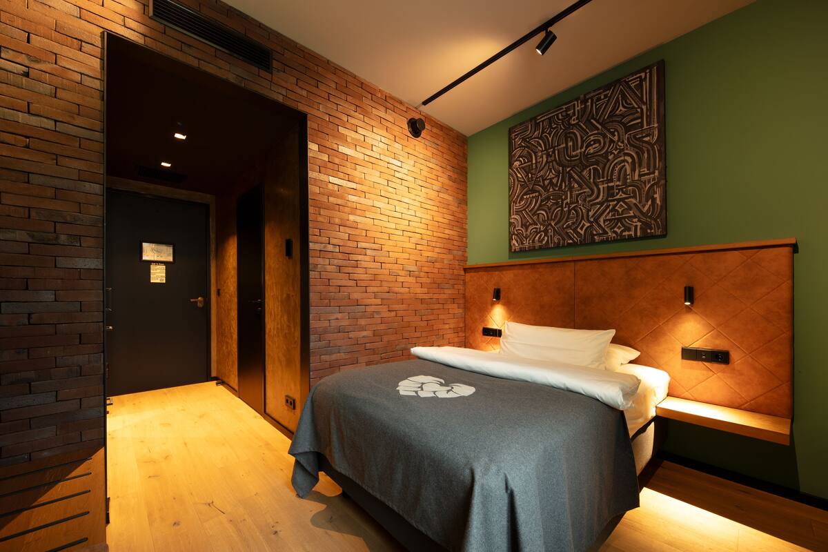 A hotel room with a brick wall, a bed with a grey bedspread, and a green accent wall with a decorative piece.