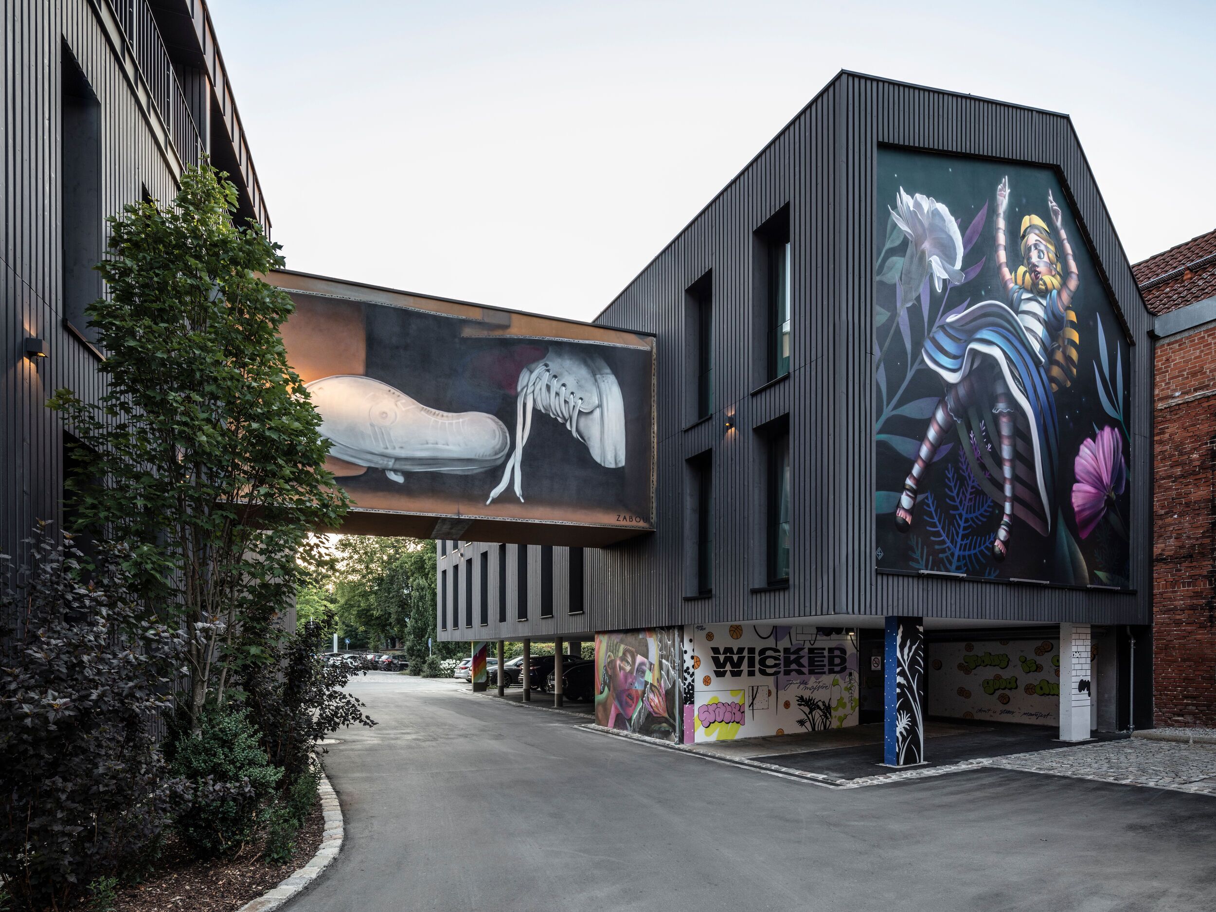 Un bâtiment moderne orné d’une grande fresque sur un de ses murs, avec un espace de stationnement et un arbre au premier plan.