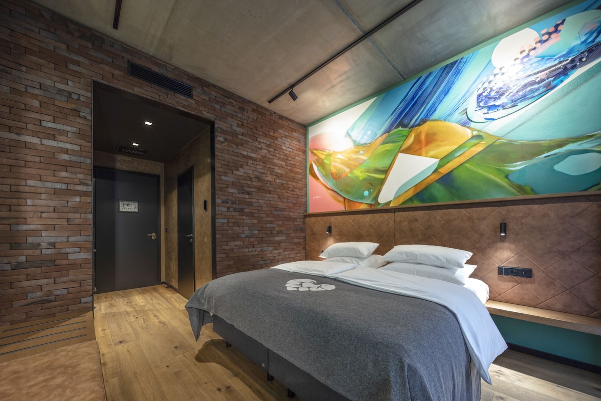 A modern hotel room with a brick wall, a large abstract mural, a wooden floor, and a bed with a grey blanket.