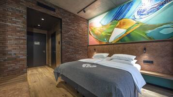 Habitación de hotel moderna con pared de ladrillo, un gran mural abstracto, suelo de madera y una cama con manta gris.