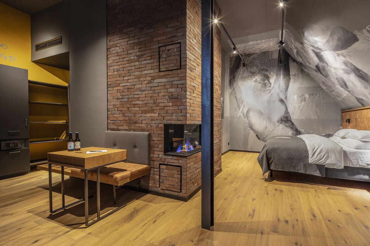 A modern room with a brick fireplace, a bed with a headboard, a small table with bottles, and a wall mural.