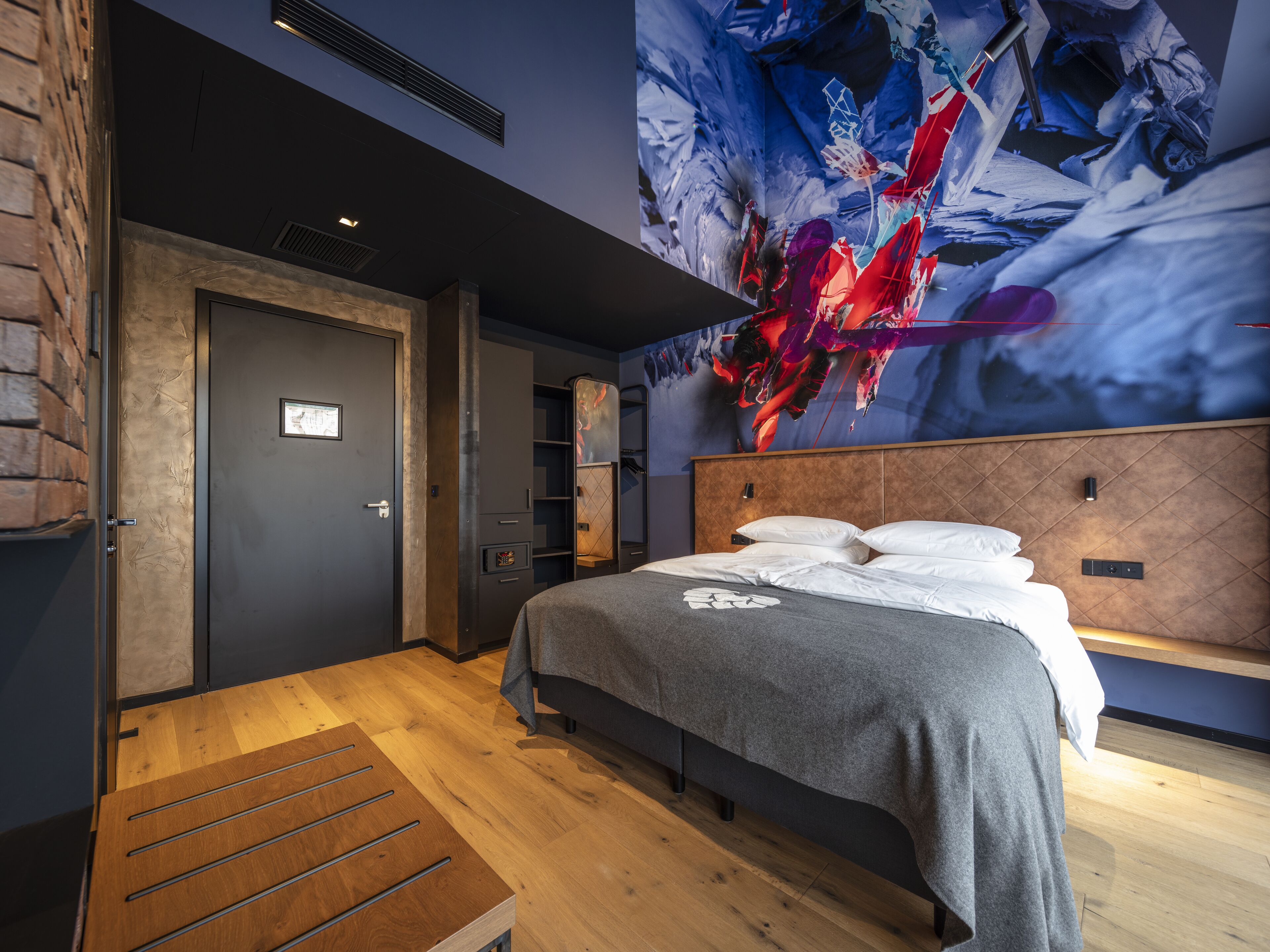 Una habitación de hotel moderna con una cama grande, piso de madera, pared de ladrillo y un cuadro abstracto de gran tamaño en la pared.