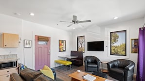 Una sala moderna con un sofá, una mesa de centro y sillas, un comedor con un banco, y una cocina con un microondas.